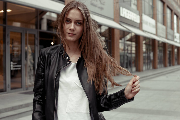 lifestyle portrait of trendy young woman with long blonde hair. Female model in black leather jacket and white t-shirt standing on the middle of the street, looking at the camera and playing hair - Photo, Image