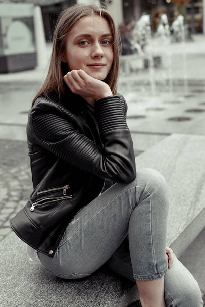 Pretty girl with blonde long hair with smiling face looking at the camera. Female model wearing black leather jacket and grey jeans. Portrait on a blurred city squares and fontaine - Zdjęcie, obraz