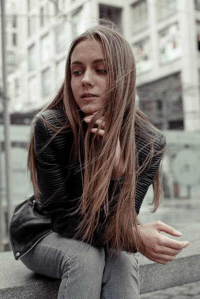 Sedící mladá žena přemýšlí. Modelka v černé kožené bundě. Dívka s rovnými blond vlasy - Fotografie, Obrázek