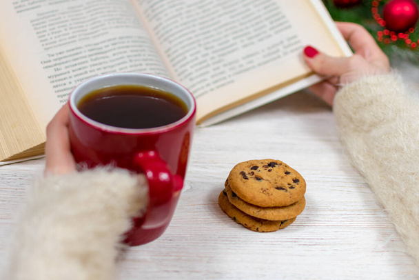 Ahşap bir masanın üzerinde bir fincan çay ve kurabiyeyle Noel ağacıyla süslenmiş kadın elleri bir kitap tutuyor. Arka plan bulanık. Kısmi odaklanma. Noel ve Yeni Yıl kavramı. - Fotoğraf, Görsel