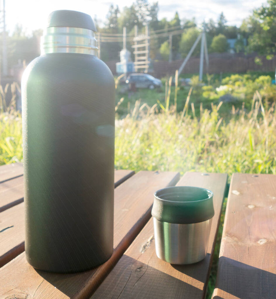 Fekete termosz és egy bögre forró teával egy faasztalon egy vidéki nyaraláson. a szabadtéri kikapcsolódás fogalma, - Fotó, kép