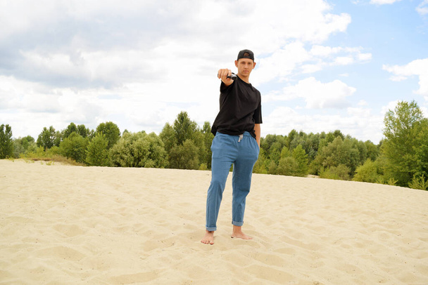 Молодой человек в черной футболке стреляет пистолетом, стоя на песке - Фото, изображение