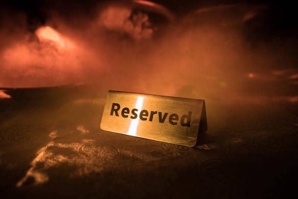 Reserved sign on the table. A tag of reservation placed on the wood table. Metal tag with reservation on dark. Reserved table in a restaurant. Creative concept with colorful lights. Selective focus - Photo, Image