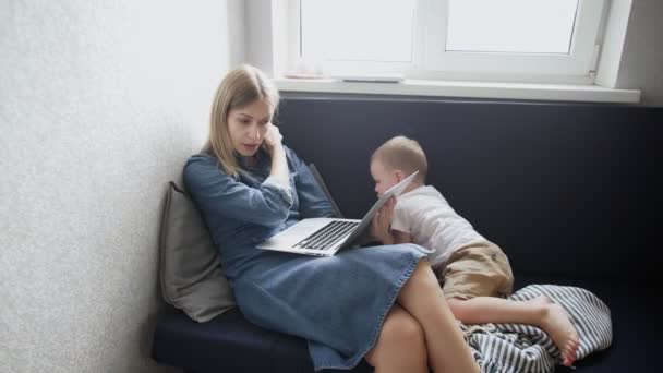 Een jonge vrouw die thuis werkt met een klein kind. Moeder is bezig met hotebook terwijl haar zoon probeert te spelen met haar - Video