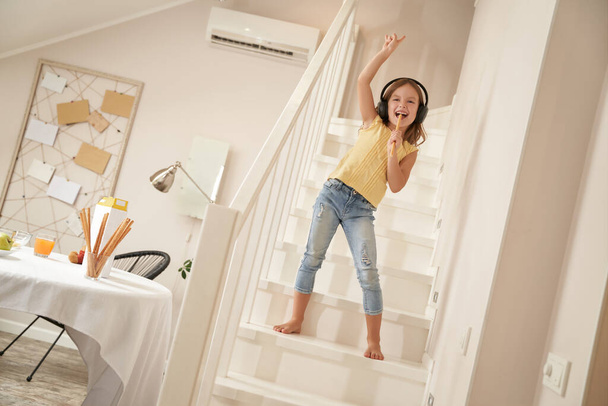 Little singer. Cute and happy caucasian girl wearing headphones standing on stairs and listening music, she is singing and having fun at home - Photo, Image