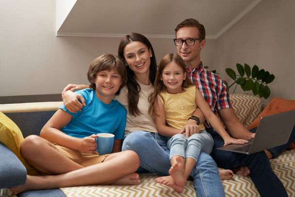 Fröhliche junge Eltern und ihre Kinder sitzen auf dem Sofa im Wohnzimmer und lächeln in die Kamera, während sie gemeinsam auf dem Laptop Filme gucken - Foto, Bild