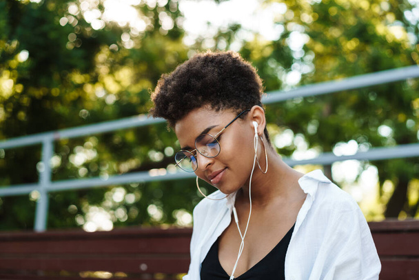 Photo of pleased african woman sitting outdoors in park while listening music - Foto, Imagen