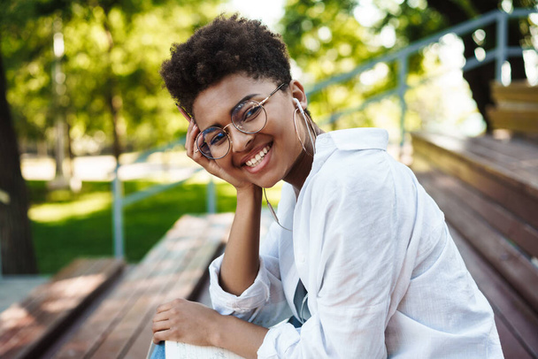 Portrait of happy smiling african positive woman sitting outdoors in park while listening music with earphones - Foto, Imagen