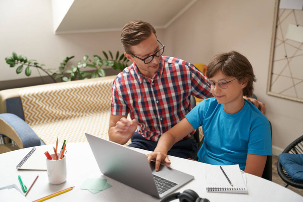 Genç baba gözlük takıyor oğluna ödevlerinde yardım ediyor, çocuk dizüstü bilgisayar kullanıyor. - Fotoğraf, Görsel