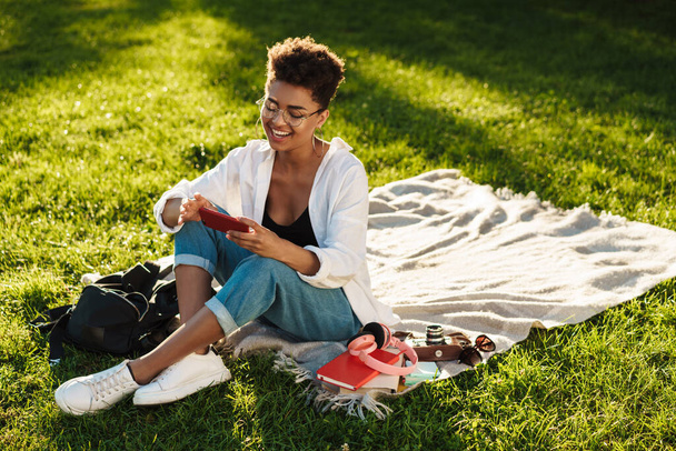 Photo of happy african woman sitting outdoors in park on a grass and using mobile phone - Fotografie, Obrázek