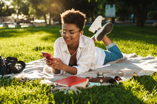 Image of positive african woman lying outdoors in park on a grass and using mobile phone while listening music - Foto, Imagem