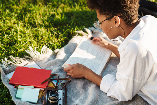 Image of concentrated african woman lying outdoors in park on a grass and reading a book - Foto, afbeelding