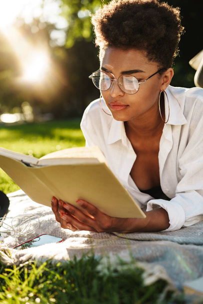 Photo of serious african woman lying outdoors in park on a grass and reading a book - Foto, immagini