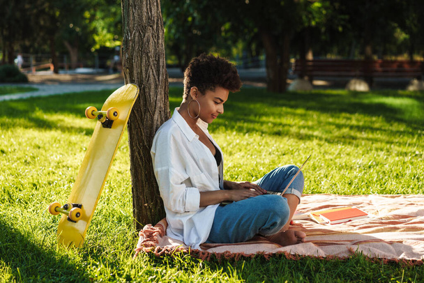 Fénykép koncentrált afrikai nő ül a szabadban a parkban egy füvet, és a laptop számítógép - Fotó, kép