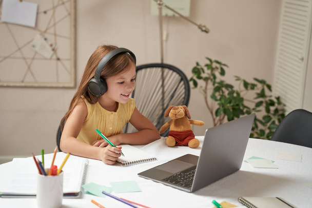 System e-learningowy. Mądra mała biała dziewczyna w słuchawkach siedzi przy stole, używa laptopa i ogląda lekcje wideo - Zdjęcie, obraz