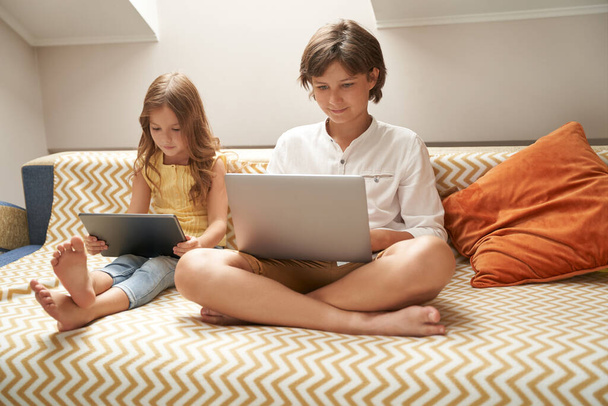 Küçük çocuklar, tatlı erkek ve kız kardeşler kanepede oturup dizüstü bilgisayar ve dijital tablet kullanıyorlar, çizgi film izliyorlar ya da evde birlikte vakit geçirirken oyun oynuyorlar. - Fotoğraf, Görsel