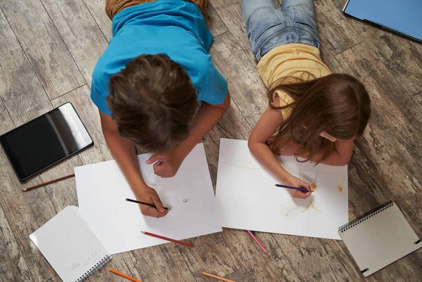 Siblings playing together at home. Top view of little boy and girl lying on the wooden floor and drawing on white sheets of paper with colorful pencils - Foto, Imagem