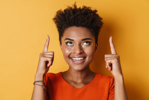 Image of cheerful african american woman pointing fingers upward and smiling isolated yellow background - Foto, afbeelding