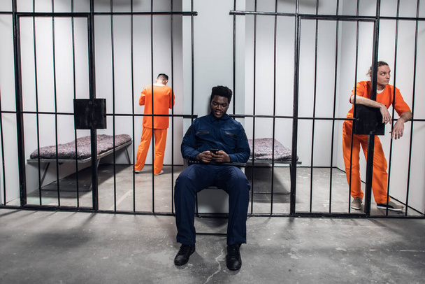 Ένας δεσμοφύλακας φυλάσσει κελιά και σερφάρει στο διαδίκτυο μέσω smartphone με επικίνδυνους κρατούμενους με πορτοκαλί στολές φυλακής.. - Φωτογραφία, εικόνα