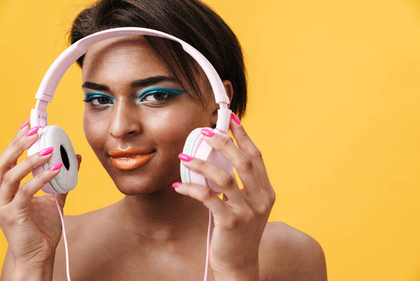 Image of shirtless african american woman with makeup using headphones isolated over yellow background - Fotografie, Obrázek