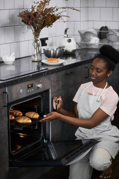 black female opens the oven, takes out cooked baked croissants - Φωτογραφία, εικόνα