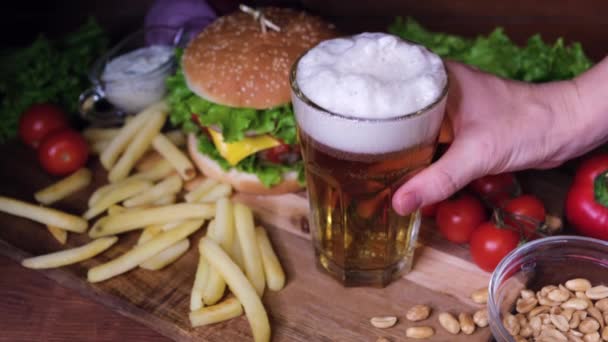 Bira, hamburger ve patates kızartmalı ahşap bir masa. El bira bardağını kaldırır ve boş olarak geri getirir. Fast food ve bira konsepti. - Video, Çekim