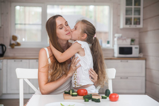 Happy family mother and child posing at home. Beautiful young mom and little daughter having fun and preparing vegetables for salad in a white kitchen in a Scandinavian style interior. Healthy food - Foto, Bild