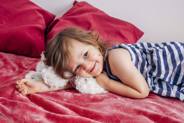 Ένα τρίχρονο κοριτσάκι ξαπλώνει στην κρεβατοκάμαρα σε ένα ροζ κρεβάτι και γελάει. - Φωτογραφία, εικόνα