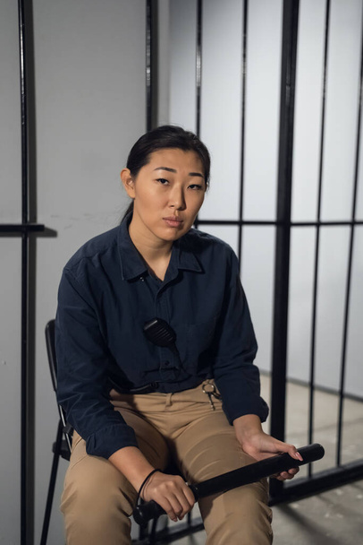 Eine junge Aufseherin in einem Gefängnis neben den Gittern mit einem Polizeiknüppel in der Hand - Foto, Bild