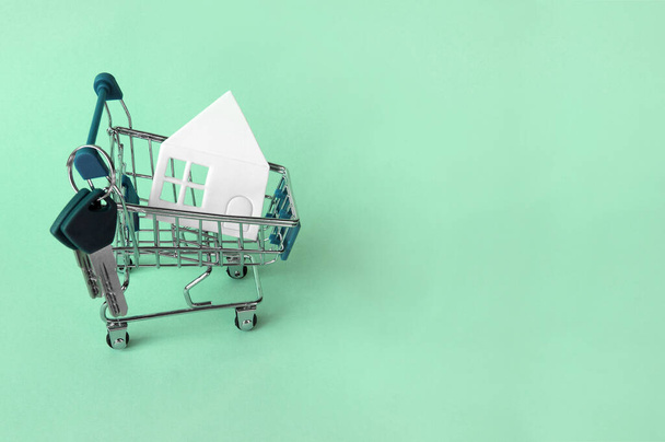 Mini winkelwagentje met een wit huis en sleutels op een groene achtergrond. Het concept van het kopen of huren van een huis, appartement, huisje en meer. - Foto, afbeelding