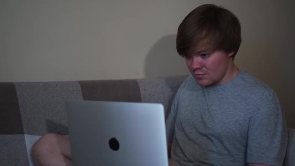 Красивий молодий чоловік сидить на дивані і використовує ноутбук у своїй кімнаті. Поняття. Блондинка в сірій сорочці працює вдома, концепція самоізоляції та фрілансу
. - Кадри, відео