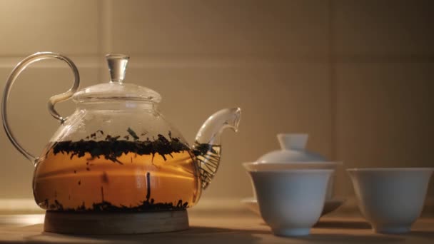 Чайник со свежими чайными листьями в горячей воде и маленькие чашки на кухонном столе. Концепция. Закрыть стеклянный прозрачный чайник с пивоварением черный чай, концепция чайной церемонии. - Кадры, видео