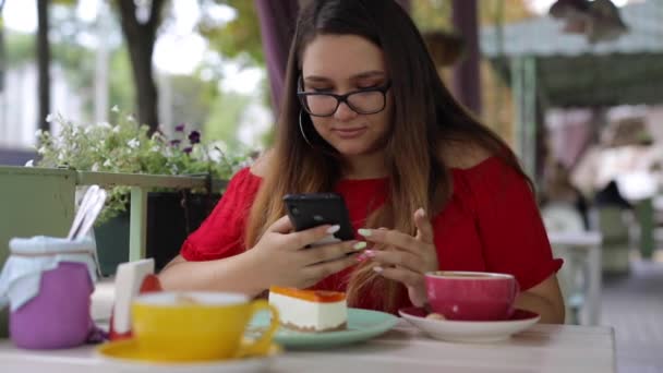 Söpö tyttö kommunikoi älypuhelimessa kahvilan pöydässä - Materiaali, video