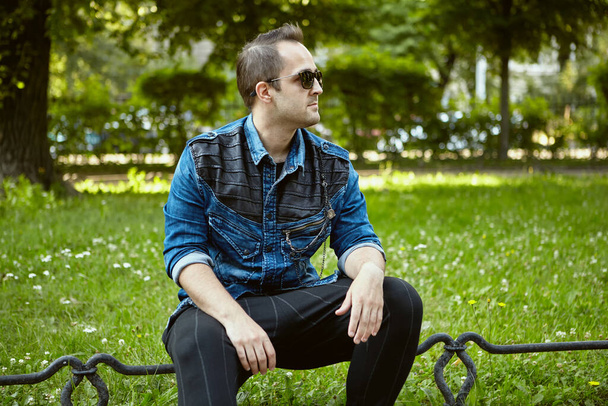 Russian man 27 years old is sitting on the lawn fence in the public park. Young male in denim jacket and sunglasses has a rest during walk over city. - Zdjęcie, obraz