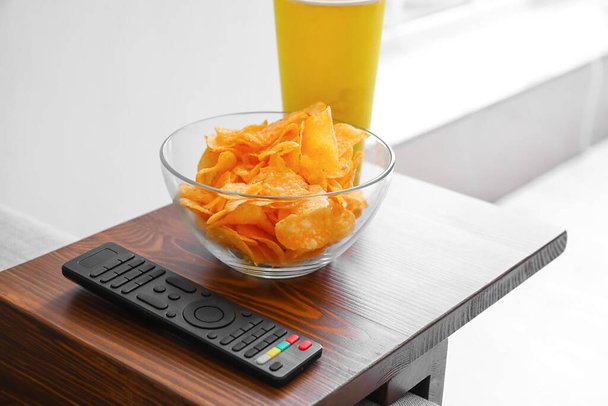 Bowl with chips and remote control on armrest table in room - Photo, Image