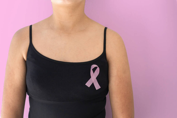 Λεπτομέρεια γυναίκας σε μαύρη μπλούζα με κορδέλα ευαισθητοποίησης για τον καρκίνο του μαστού σε ροζ φόντο. Σύμβολο πρόληψης - Φωτογραφία, εικόνα