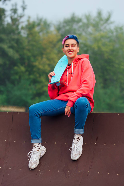 Young girl with short multicolored hair and nose piercing dressed in pink hoodie sitting with skateboard in skatepark during morning fog. Portrait of a tomboy with plastic skateboard - Фото, зображення