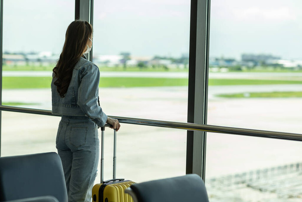 Asian young traveler woman at airport wearing face mask standing with luggage looking the airport gate windows at planes on airport runway due to coronavirus or COVID-19 pandemic - 写真・画像