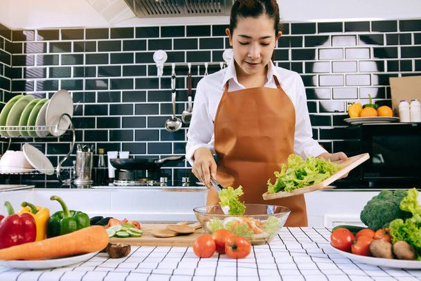 Femme au foyer asiatique utilise des pinces pour prendre la salade sur la planche à découper en bois sur la tasse à salade dans la salle de cuisine. - Photo, image
