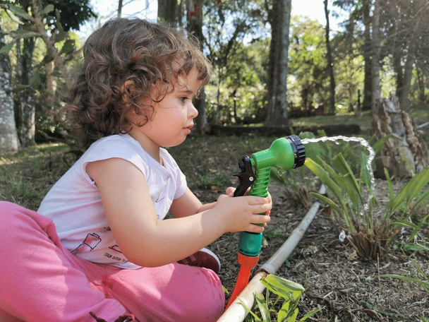 Χαριτωμένο Βραζιλιάνικο κοριτσάκι που διασκεδάζει παίζοντας με το λάστιχο νερού στον κήπο. - Φωτογραφία, εικόνα