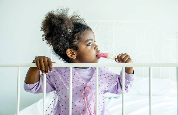 Αφρικανική αμερικανική χαριτωμένο κοριτσάκι κρατώντας τρώει παγωτό στο κρεβάτι στην κρεβατοκάμαρα - Φωτογραφία, εικόνα