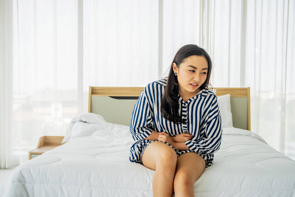 Nešťastná mladá asijská žena trpí bolestí břicha, zatímco sedí doma na posteli - Fotografie, Obrázek