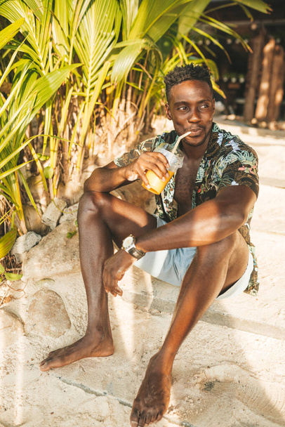 Portrait of african american man wearing a hawaiian shirt drinking mango shake in outdoor cafe - 写真・画像