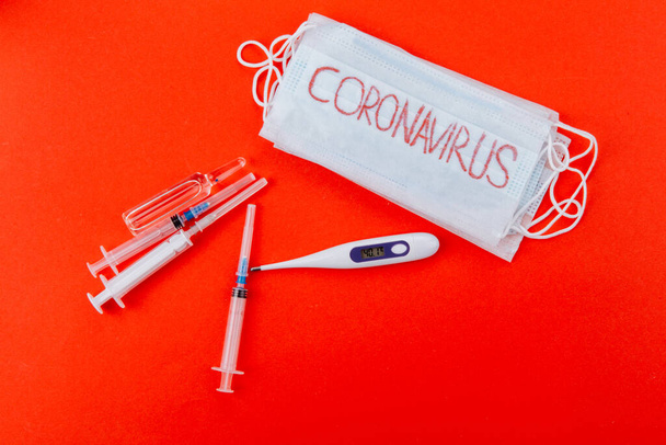 Protective medical mask with the inscription coronavirus, syringes, ampoules and thermometer on a red background. Protective mask as a precaution for the spread of Covid-19 coronavirus worldwide. - Fotografie, Obrázek