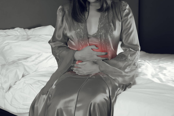 Egy szatén hálóinges nő, aki hasfájástól szenved, miközben az ágyban ül éjszaka. - Fotó, kép