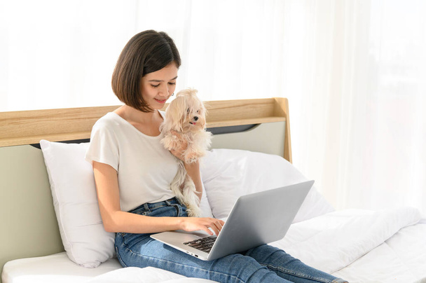 Krásná mladá asijská žena pracující na posteli v ložnici doma se svým maltským psem, veselý a pěkný pár s lidmi a domácím mazlíčkem - Fotografie, Obrázek