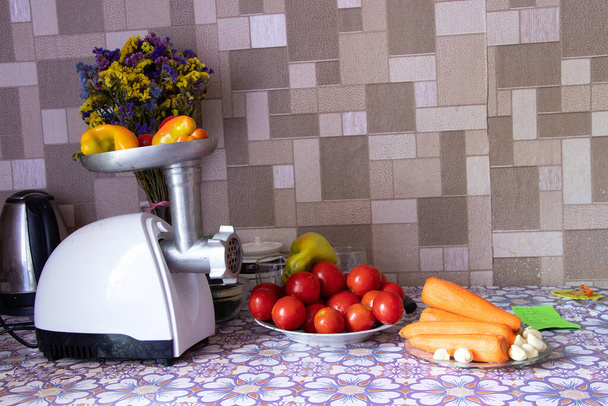 электрическая мясорубка с перцем на столе рядом с помидорами и морковью на столе на кухне, приготовление домашней адиджики, домашние приготовления - Фото, изображение