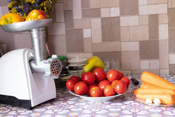 elektrische Fleischmühle mit Pfeffer auf dem Tisch neben Tomaten und Karotten auf dem Tisch in der Küche, Kochen hausgemachte adijika, hausgemachte Zubereitungen - Foto, Bild