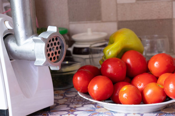 elektryczna szlifierka do mięsa z pieprzem na stole obok pomidorów i marchwi na stole w kuchni, domowej roboty adijika, domowe przetwory - Zdjęcie, obraz