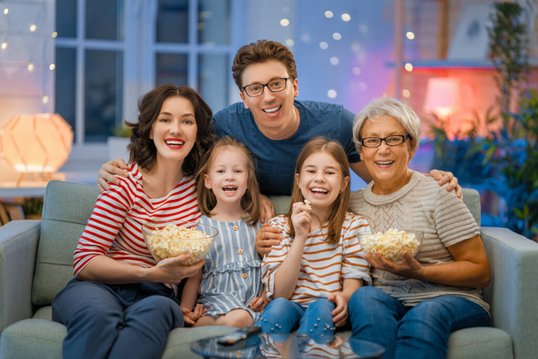Happy family watching projector, TV, movies with popcorn in the evening at home. Granny, mother, father and daughters spending time together. - 写真・画像